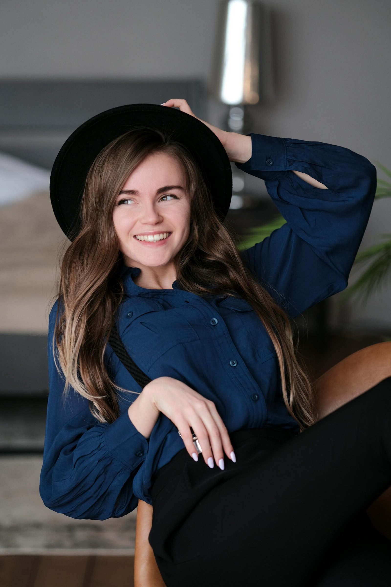 Woman with long brown hair and hat smiling while sitting indoors, exuding a warm and relaxed vibe.